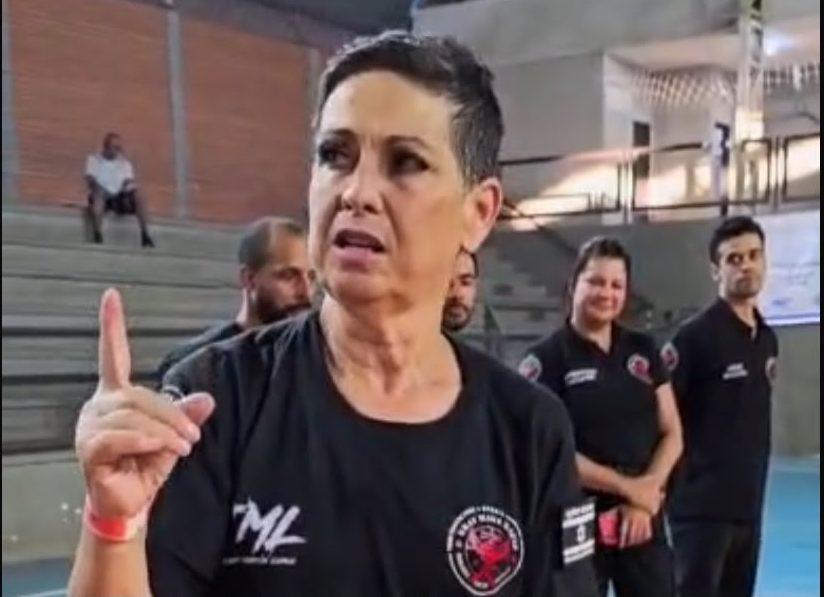 Mulher idosa pratica golpe de defesa pessoal em treino de Krav Maga ao lado de instrutor, em academia.