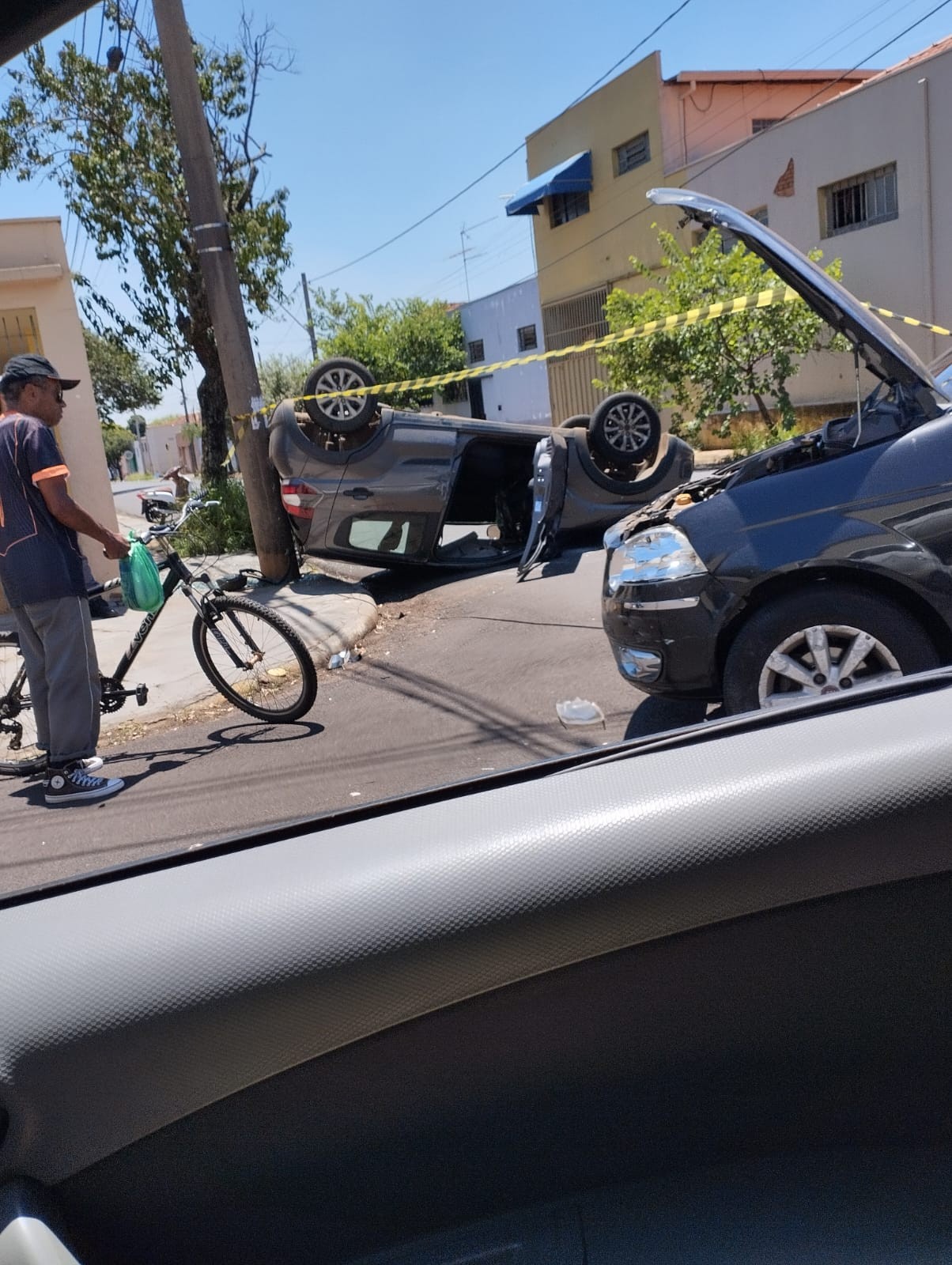 Carro cinza de rodas para o alto junto a um poste, com a área isolada por fita e um carro preto de capô aberto em primeiro plano