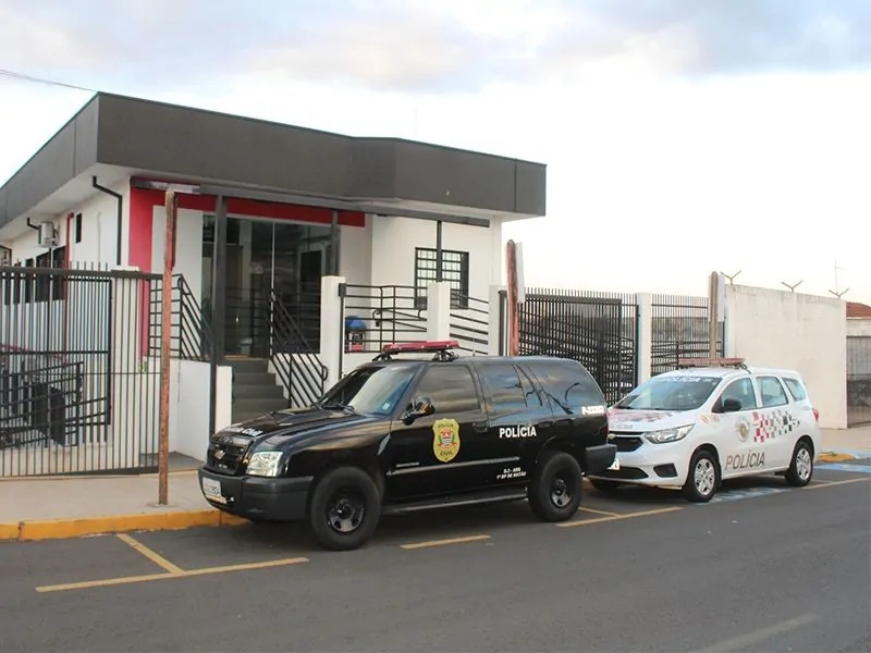 Viatura preta da Polícia Civil e viatura branca da Polícia Militar estacionadas em frente a um prédio de delegacia