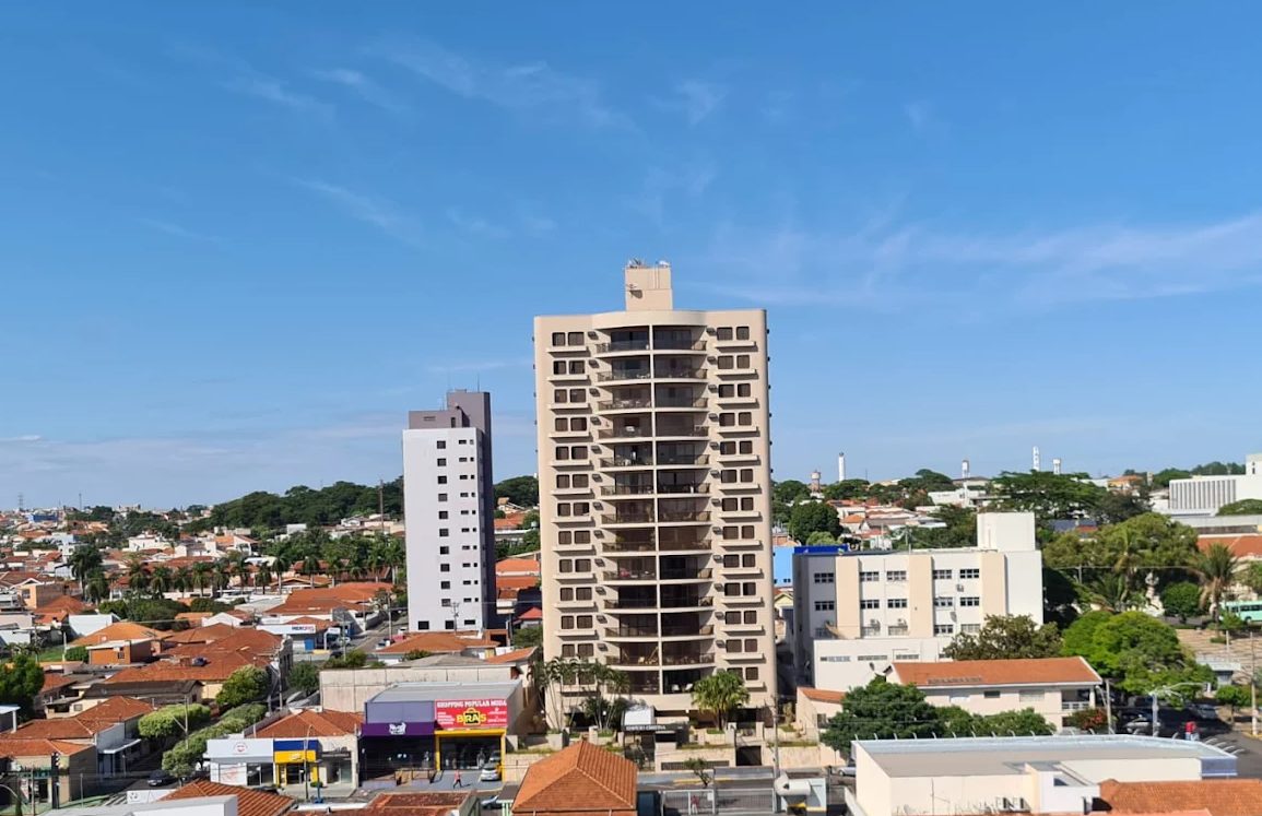 Céu azul e sol sobre uma cidade do interior paulista, com prédios baixos, árvores e uma praça ao fundo.