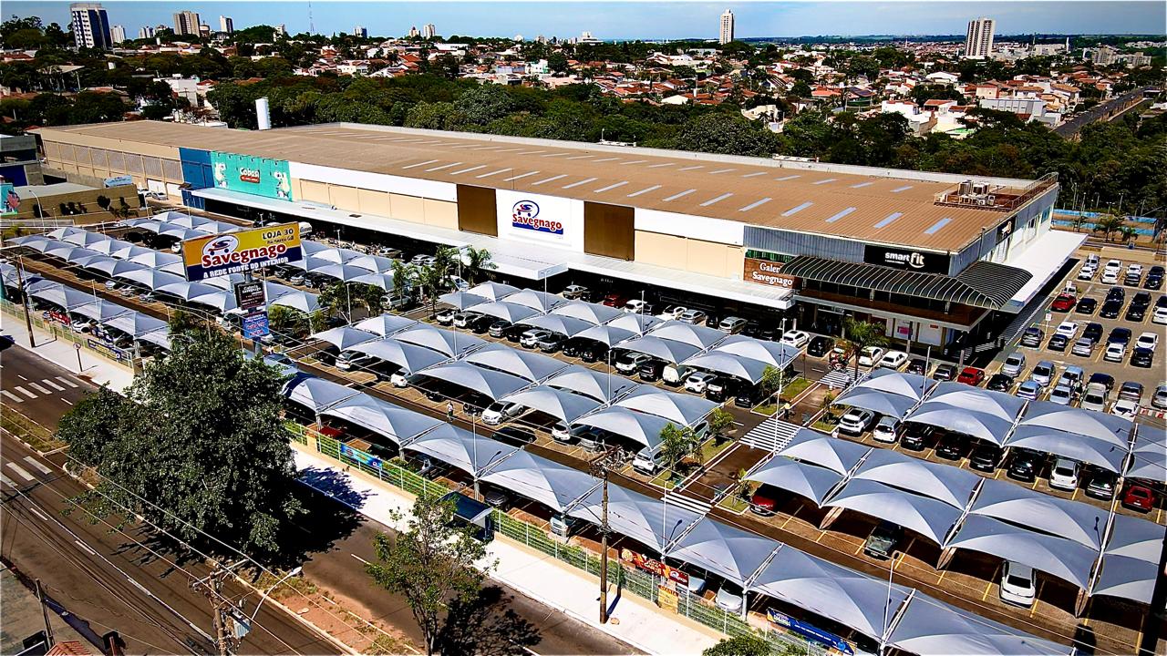 Vista aérea de um supermercado grande com estacionamento coberto por toldos claros e a cidade ao fundo